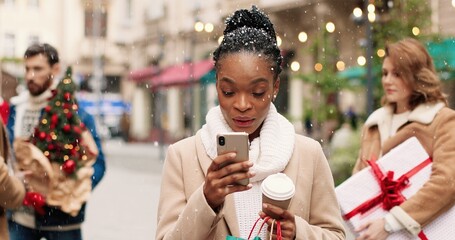 Joyful African American woman in decorated city with cup of coffee and texting on smartphone and receiving holiday greetings. Close up portrait of female with bags after shopping typing on cellphone