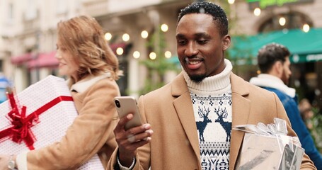 Joyful African American man in decorated crowded city with xmas gift box texting on smartphone and sending holiday greetings. Close up portrait of male with present after shopping typing on cellphone