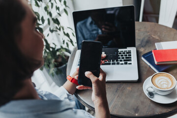 Young woman sitting in coffee shop at wooden table, drinking coffee and using smartphone.On table is laptop. Girl browsing internet, chatting, blogging. Female holding phone and looking on his screen.