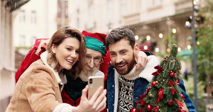 Close up portrait of happy Caucasian female and male standing with Santa Claus and taking selfie pictures on street. Joyful couple with Santa recording video for vlog. Christmas spirit concept