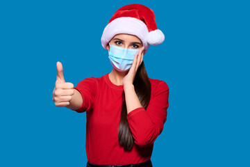Fototapeta na wymiar Excited and shocked woman wearing Santa hat and face protective medical mask showing thumb up gesture, blue isolated background.