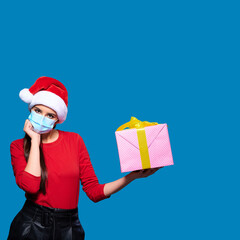 Upset woman wearing Santa hat and face protective medical mask and touching face, blue isolated background.