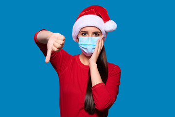 Excited and shocked woman wearing Santa hat and face protective medical mask showing thumb down gesture, blue isolated background.