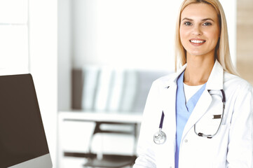 Blond female physician is standing at her workplace near desktop computer. Woman-doctor is excited and happy of her profession. Medicine concept