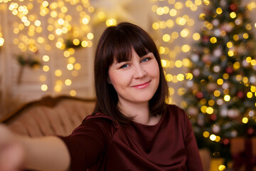 young beautiful woman taking selfie photo in decorated living room with Christmas tree and festive led lights