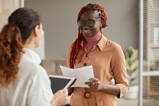 Waist up portrait of young African-American businesswoman talking to female manager and smiling cheerfully while standing in modern office interior