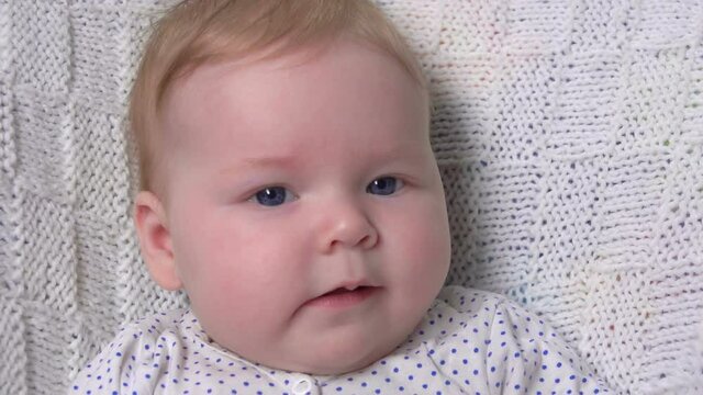 A cute little blue-eyed baby on a white knitted blanket is looking at the camera