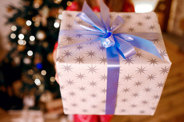 A child girl in red pajamas holds a Christmas present in her hands on the background of a New Year tree with lights. New year and christmas holiday concept. Gift box tied with blue ribbon close up