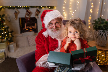 Fototapeta na wymiar Funny Santa Claus sitting on his rocker with little cute boy sitting on his knee, opening up a gift with something special together. Christmas spirit, holidays and celebrations concept
