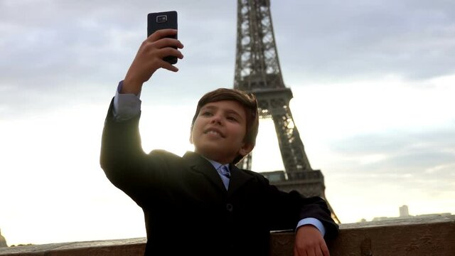 A smiling teenage boy in a suit is making a selfie on the phone on the background of the Eiffel tower at the dawn, Paris, France