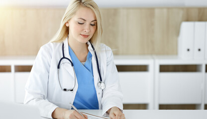 Woman-doctor at work while sitting at the desk in sunny clinic. Blonde cheerful physician filling up medication history record form