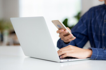 Closeup hand of young man working laptop computer and reading smartphone on internet online on desk at home, freelance male using phone with social media, business and communication concept.