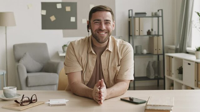 Medium close-up POV of cheerful Caucasian guy sitting by desk at home office smiling, waving hand hello, wearing wired headset, talking, looking on camera. Man having online call, working remotely