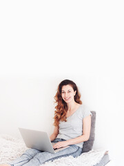 Naklejka premium Young girl with a grey laptop sitting on a bed, white space on the background. The woman has long hair, looks happy and content