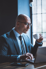 Respectable businessman drinks coffee working with computer against of window. Side view of good...