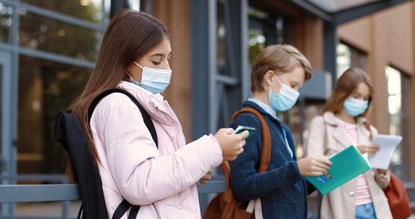 Portrait of teen Caucasian school children standing outdoors near school and reading. Concentrated beautiful girl in mask reads from copybook. Boy with planner before classes. Autumn concept