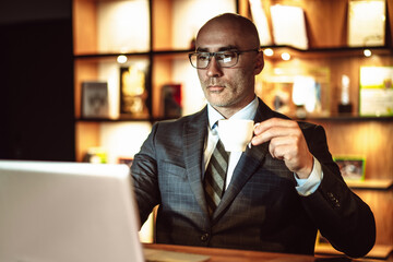 Successful business man reads worlds news. Middle aged male person looks on laptop screen drinking coffee while working in business center or hotel cafe.