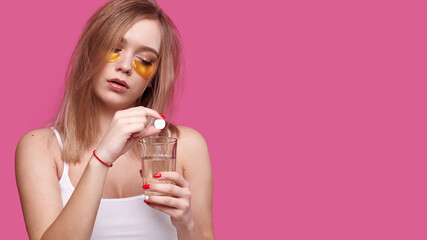 Woman with flowing hair puts pain reliever pill into glass of water after hangover on pink isolated background