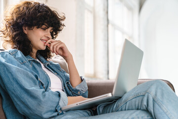 Joyful curly girl using laptop and smiling while sitting on sofa