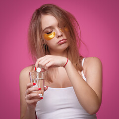Fototapeta premium Sleepy woman with flowing hair puts pain reliever pill into glass of water after hangover on pink isolated background