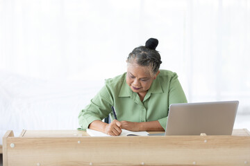 senior woman writing in notebook and using laptop computer at home