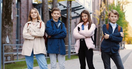 Caucasian cheerful school kids standing on street near school and posing to camera while smiling in...