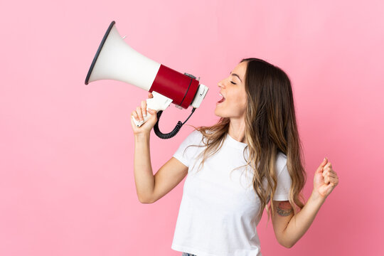 Young Romanian woman isolated on pink background shouting through a megaphone to announce something in lateral position
