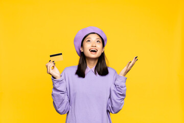 excited asian woman in beret holding credit card and looking up isolated on yellow