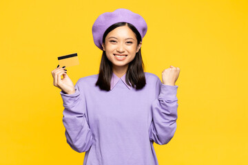 Fototapeta na wymiar excited asian woman in beret holding credit card and rejoicing isolated on yellow