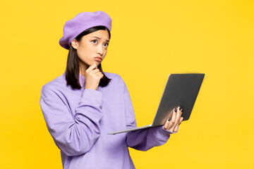 pensive asian woman in beret using laptop isolated on yellow