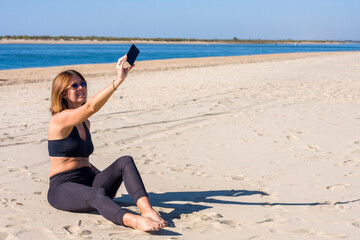 beautiful young girl sitting on the sand on the beach taking a picture with her mobile phone