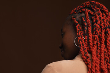 Back view portrait of stylish African-American woman with braided hair looking away while posing against dark brown background in studio
