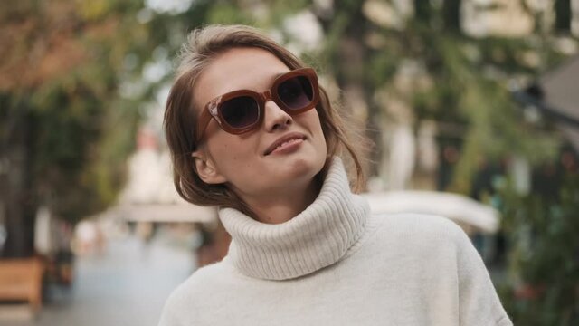 Attractive stylish girl in sunglasses dressed in white cozy sweater texting with friends during walk through city street