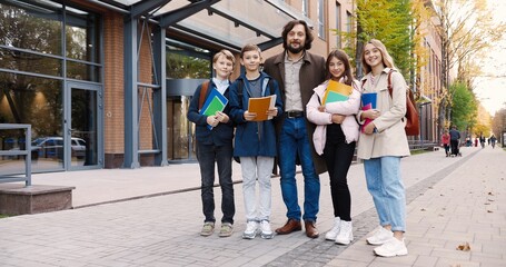 Joyful schoolboys and schoolgirls with colorful copybooks standing with man teacher on street outdoor. Happy male professor hugging his students. Caucasian girls and boys pupils near school