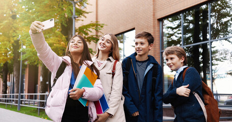 Portrait of happy school kids taking selfie photo on smartphone with friends while standing outdoors and making funny faces. Caucasian classmates taking pictures on cellphone and smiling