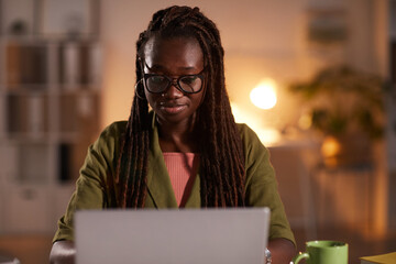 Front view portrait of young African-American woman using laptop while working in office or at home lit by cozy dim lighting, copy space © Seventyfour