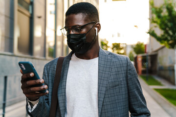 Young african american man in face mask using earphones and mobile phone