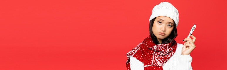 upset sick asian woman in hat and scarf holding electronic thermometer isolated on red, banner