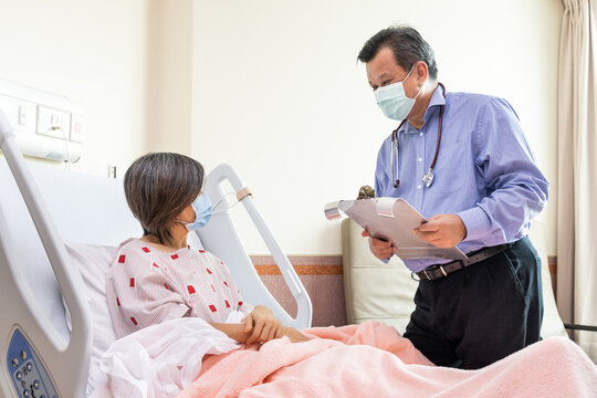 Confident Asian Doctor With Face Mask Explaning To Cheerful Female Patient On Medical Condition Lying On Bed In Hospital.