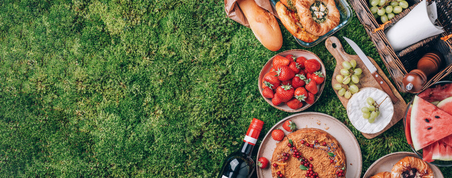 Picnic straw basket with healthy food, accessories. Summer picnic with cake, fruits, cheese, wine and snacks on plaid over green background. Top view. Copy space. Summer family lunch. Romantic picnic