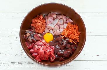 Natural raw dog food chopped ingredients in bowl on white wooden background. Meat, liver, testicles, minced salmon, chicken egg and carrot.