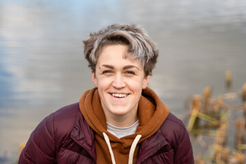 Young woman with stylish short hair, dressed in a gender neutral style against water surface. Smiling face.