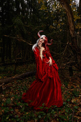 Maleficent Woman in Red Clothing and Horns in dark Forest. Posing in magik forest