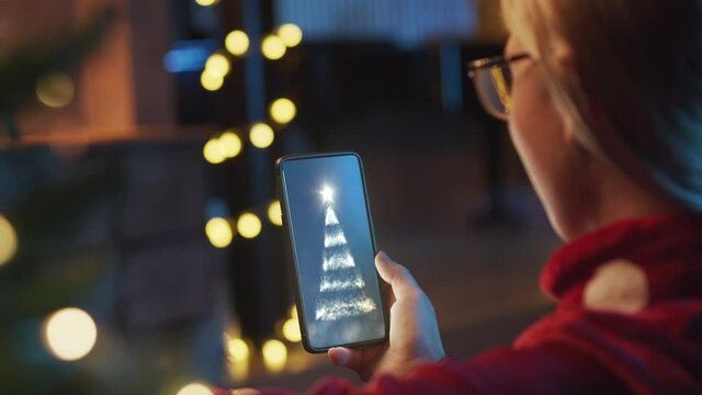 Back view of young woman holding mobile phone. Woman watching, smiling for a Christmas card from her friends, parents.In background Christmas decorations and light. Christmas outfit at home.