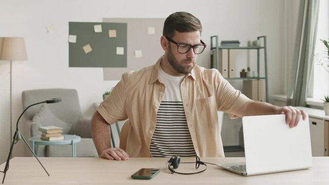 Medium of young Caucasian man wearing glasses moving to desk on chair at home office, opening laptop cover, putting headset on, and starting typing on keyboard. Person getting started for remote work