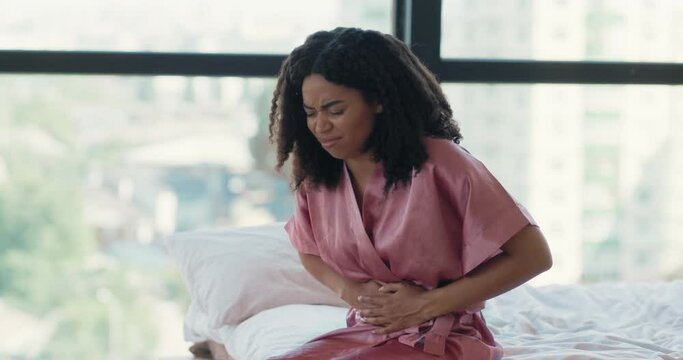 Sick black woman suffering from stomach ache sitting in bed, feeling abdominal or menstrual pain in morning