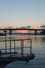 Fototapeta premium オレンジ色の夕焼けとシルエットの高架橋のある凪の港と船の先端
