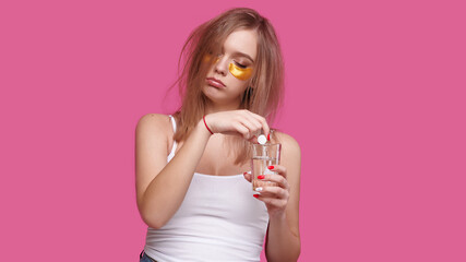 Sleepy woman with flowing hair puts pain reliever pill into glass of water after hangover on pink isolated background