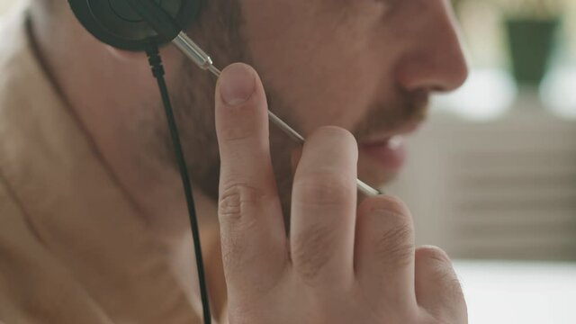 Extreme close-up of bearded unrecognizable male person below eyes part of face in profile on blurred background. Man wearing headset touching microphone part with fingers talking