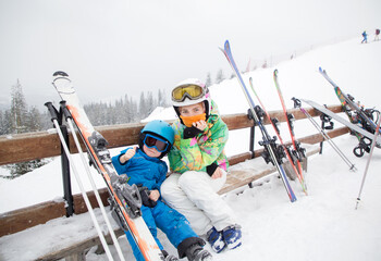 Fototapeta na wymiar toddler boy and teenage girl ,brother and sister, are resting on bench, taking break from skiing in mountains. Winter sports for family. Children's skiing lesson at alpine school. Seasonal joys
